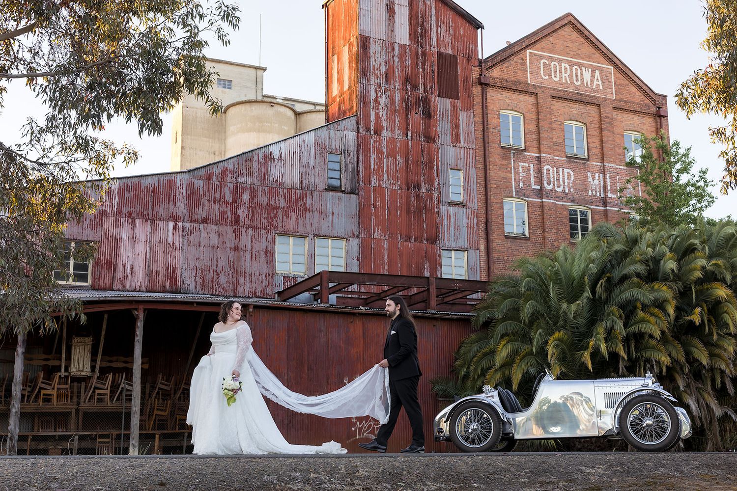 A Bride and Groom walk with the Groom holding her veil outside the Corowa Distilling Co. with a Vintage MG car in the background.