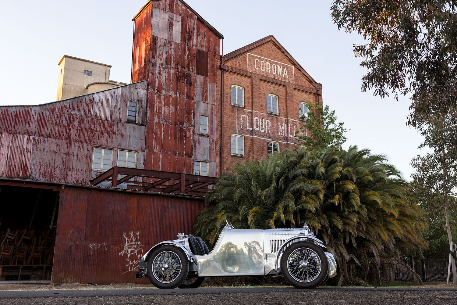 A silver vintage MG car outside the Corowa Distilling Co. building at sunset.