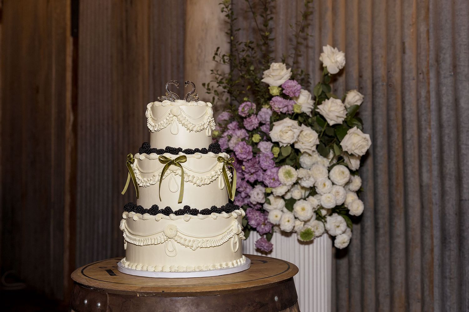 Elegant three-tier white wedding cake with delicate piping displayed next to purple and white floral arrangement.