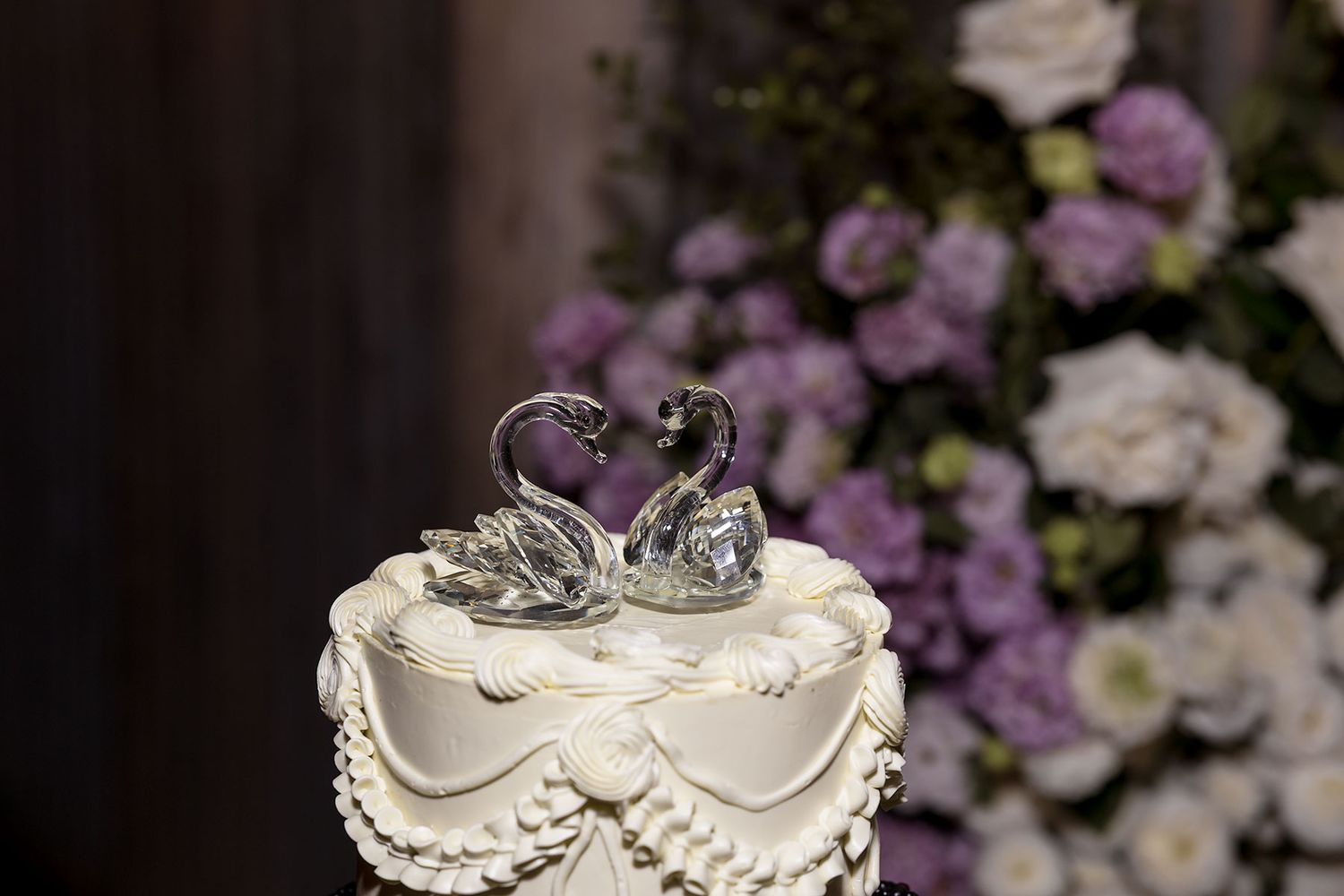 White wedding cake with crystal swans on top surrounded by purple and white floral arrangements.