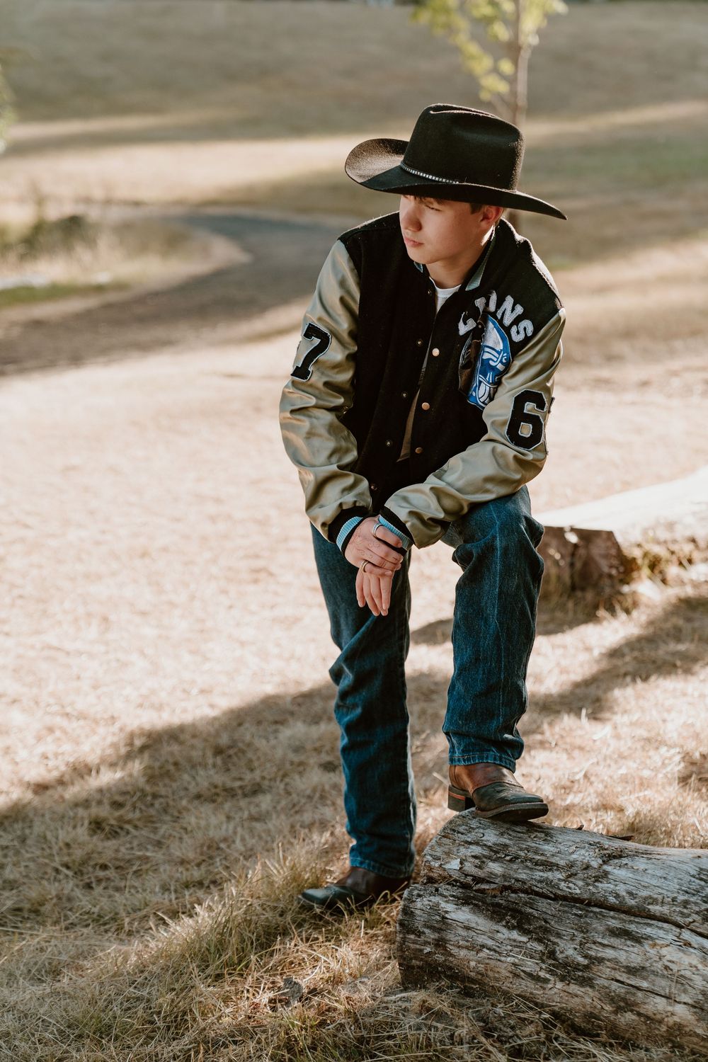 Young boy in varsity jacket and cowboy hat posing on a log in sunny field.