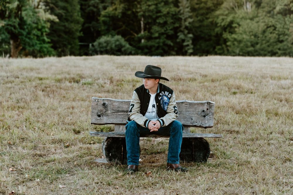 Young boy in cowboy hat and varsity jacket sitting on rustic bench in open field.