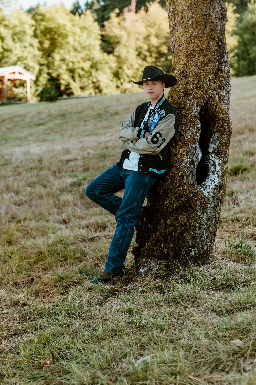 Teen boy in cowboy hat and varsity jacket leaning against a tree in a meadow.