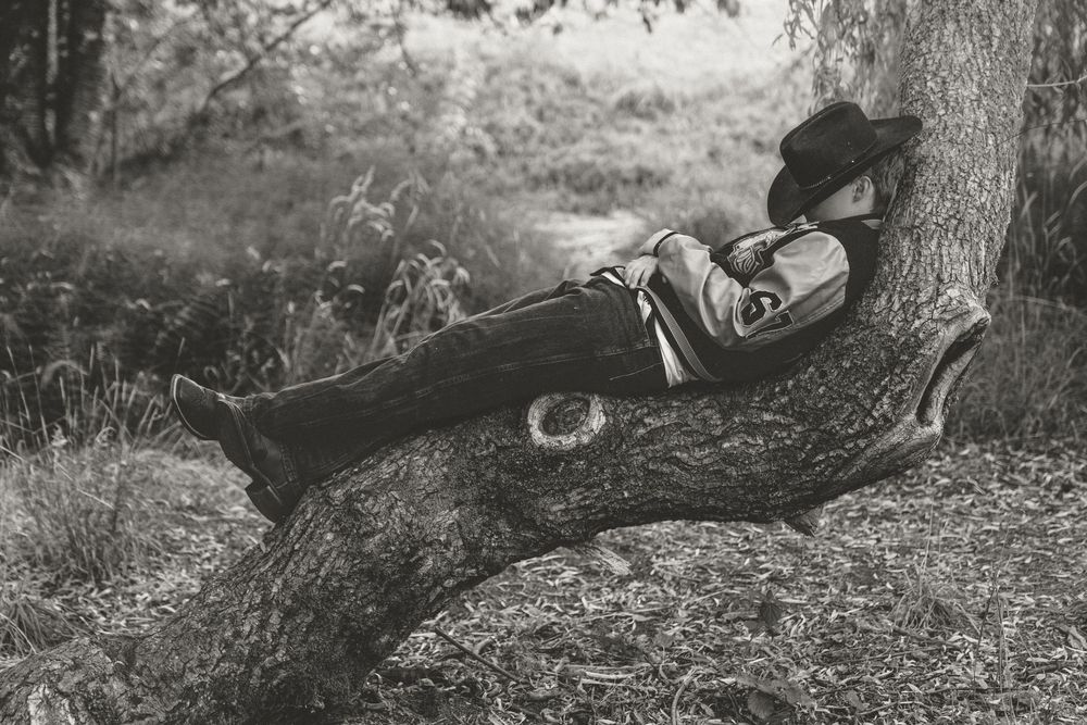 Black and white photo of a boy in cowboy hat relaxing against a tree outdoors.