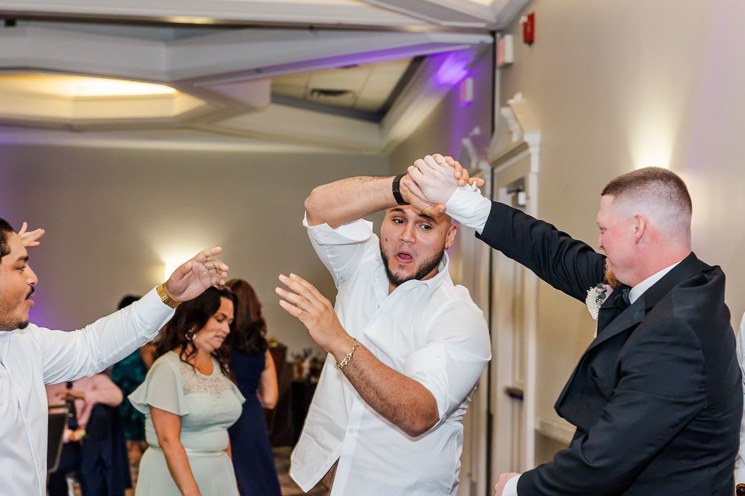 A group of people dancing and celebrating at a wedding reception with white and purple lighting.