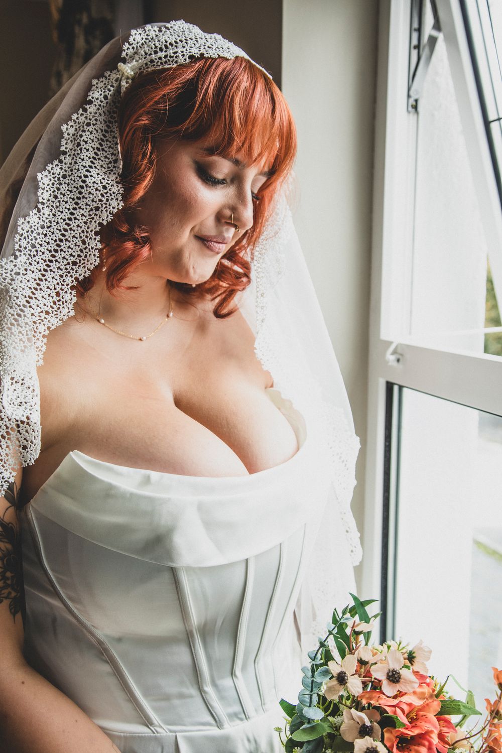 A bridal portrait showcasing an elegant white gown, delicate veil, and vibrant floral bouquet near a window.