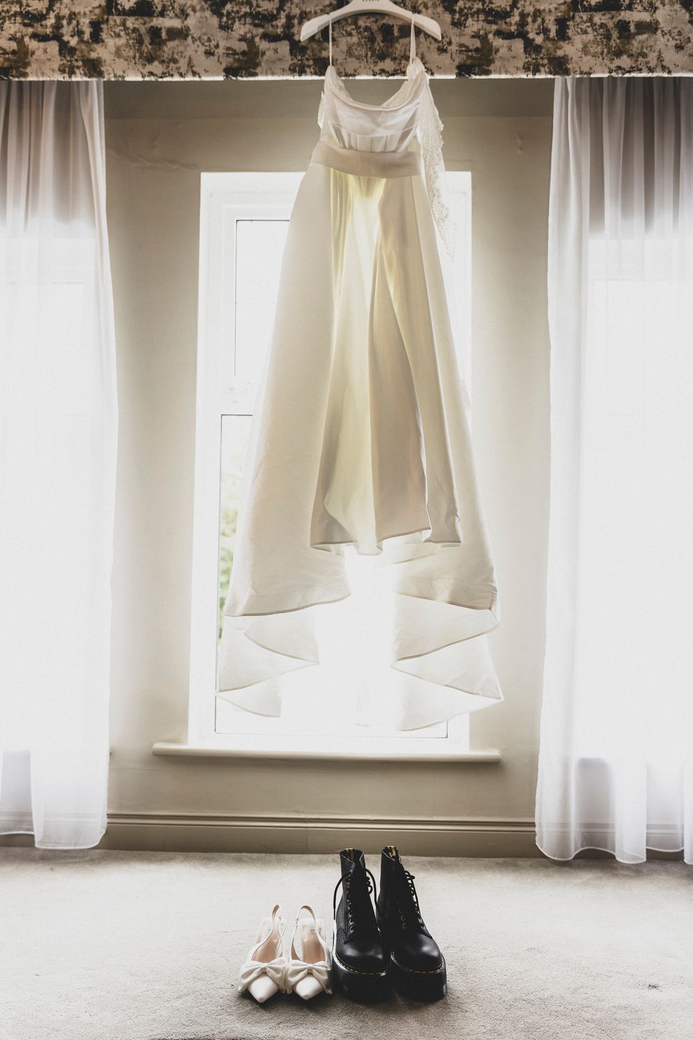Wedding dress and shoes displayed by a window with soft white curtains creating a romantic atmosphere.