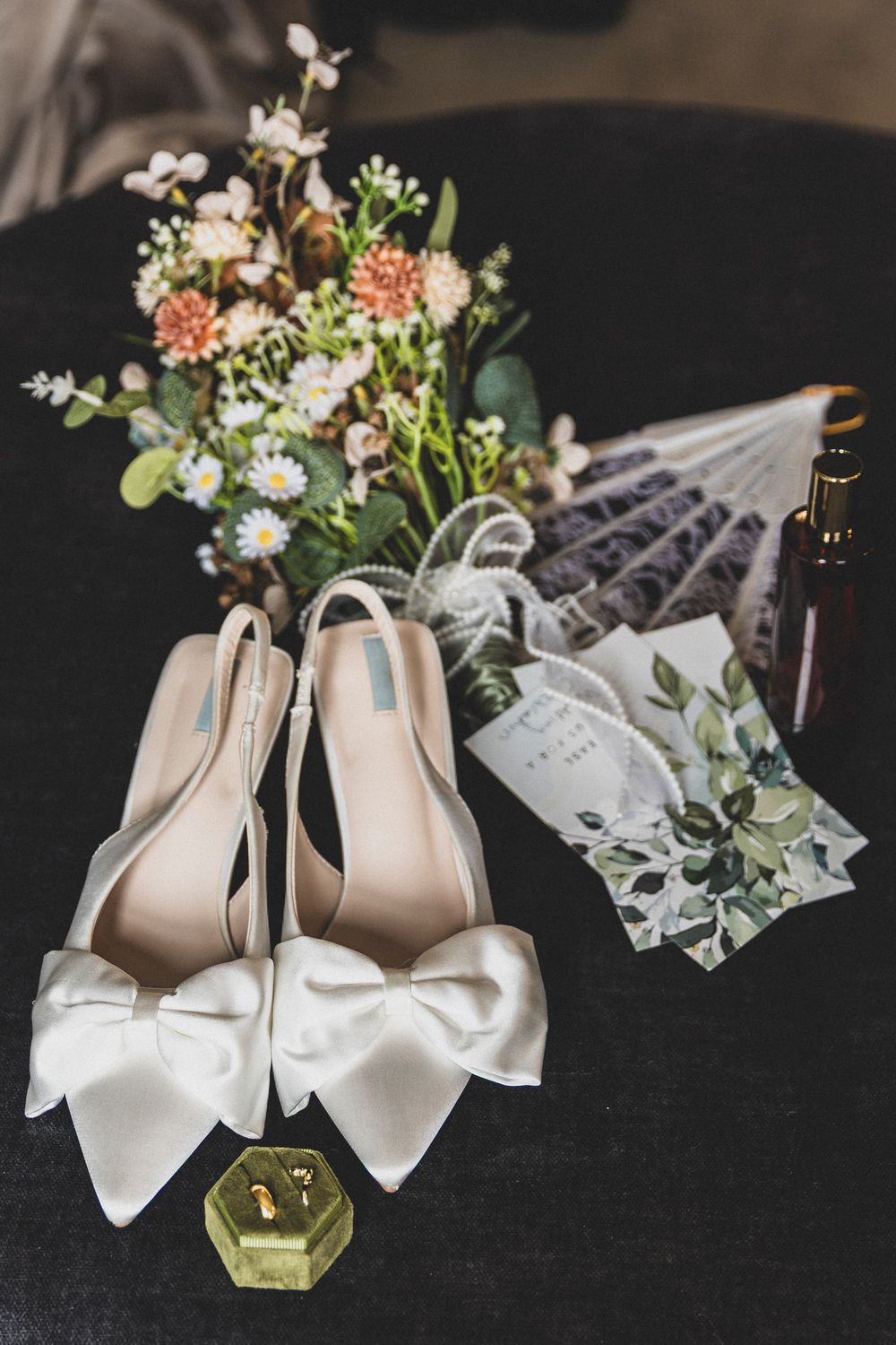 White bridal shoes with bows and floral arrangements displayed on dark surface.