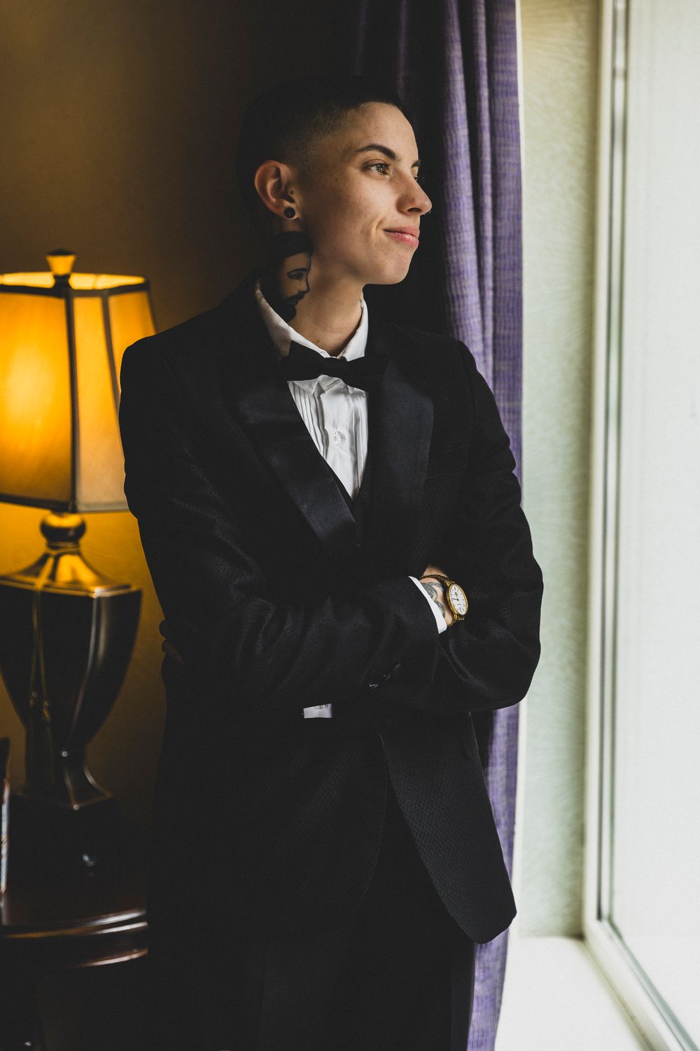A person in a black suit stands thoughtfully by a window with warm lamp light in an elegant indoor setting.