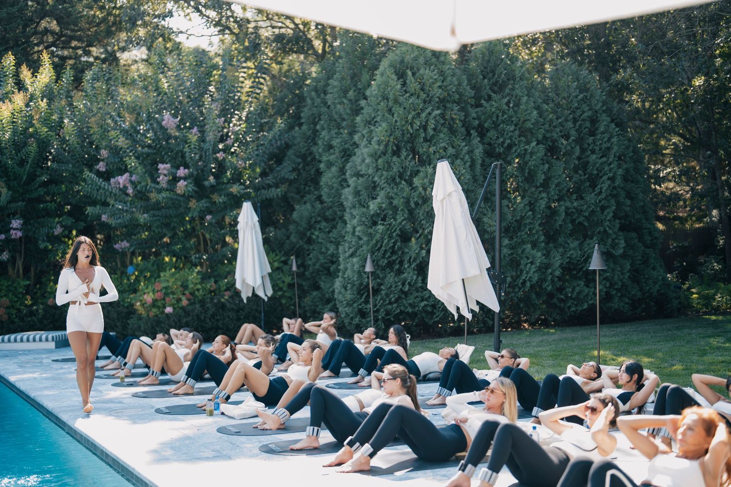 People relaxing on lounge chairs by a pool surrounded by lush greenery on a sunny summer day.
