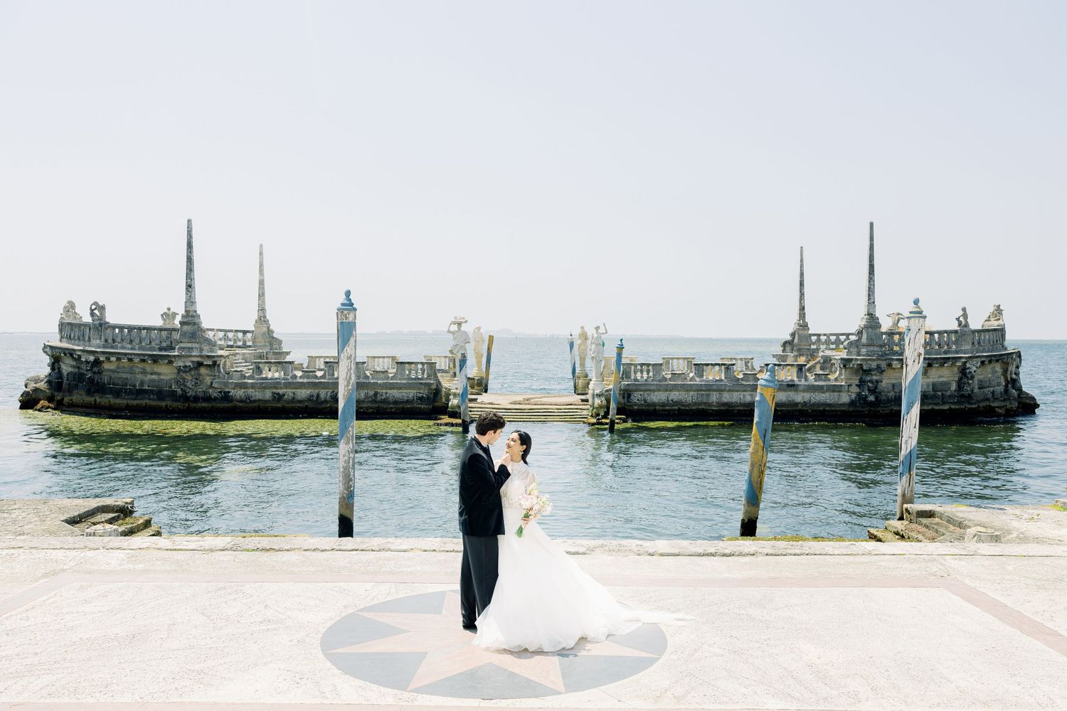 Wedding couple stands on a circular platform by the water with two large ferry boats docked in the background.