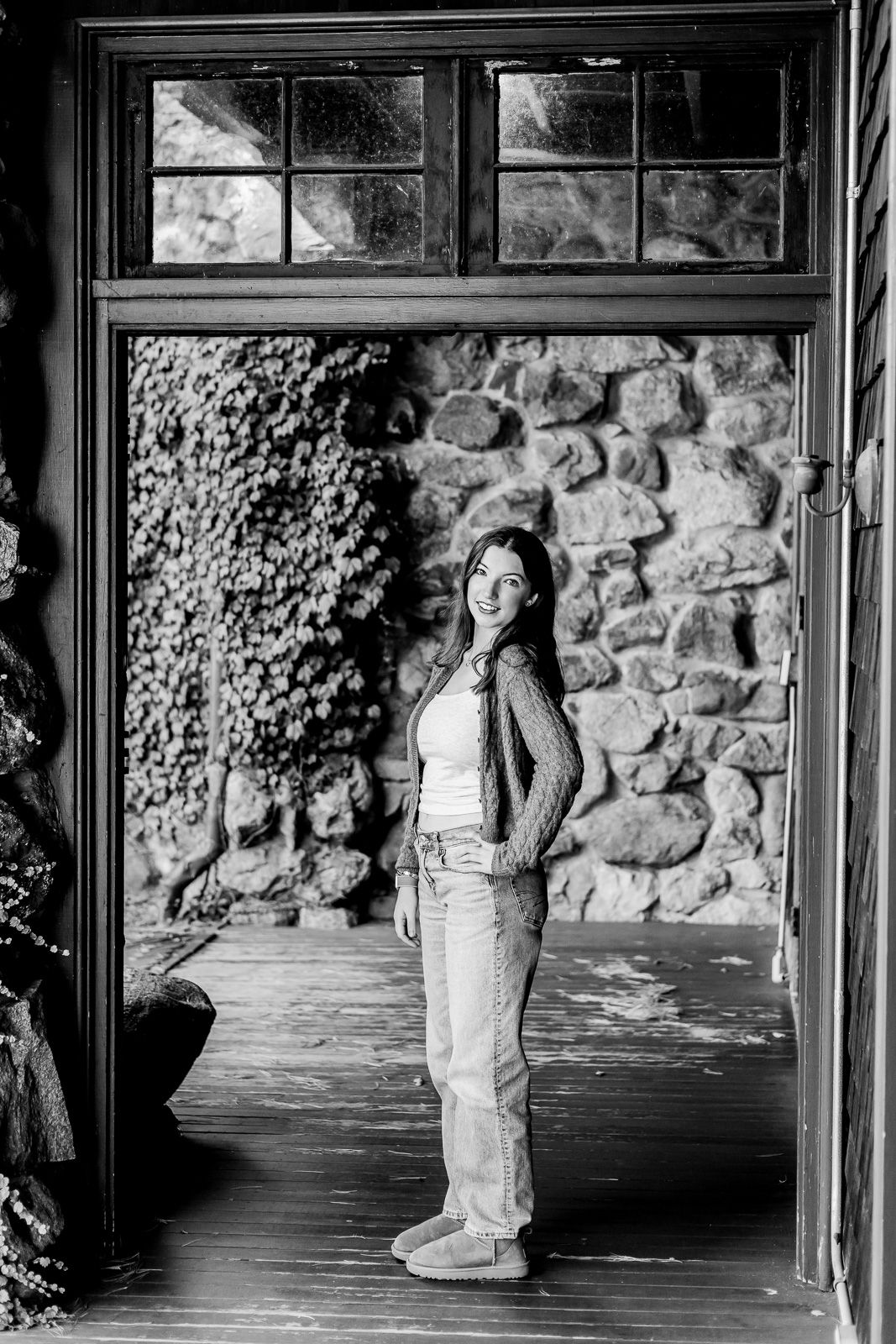 Black and white portrait of a person standing in a doorway against a rustic stone wall with climbing vines.