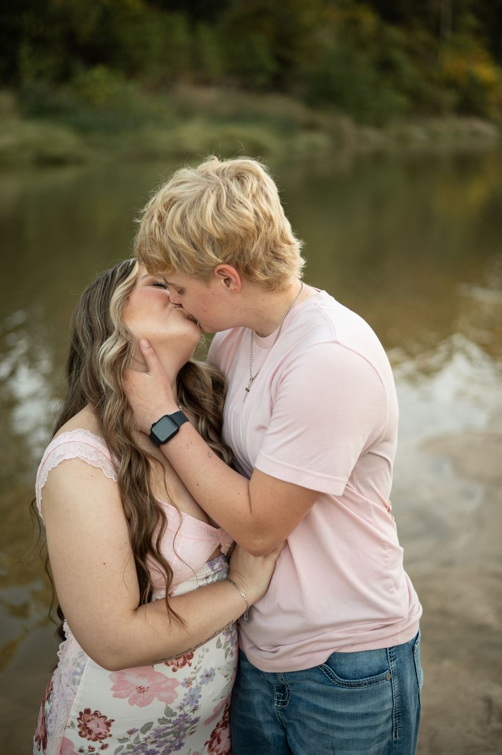 A couple shares a tender romantic moment by a serene lake with trees in the background.