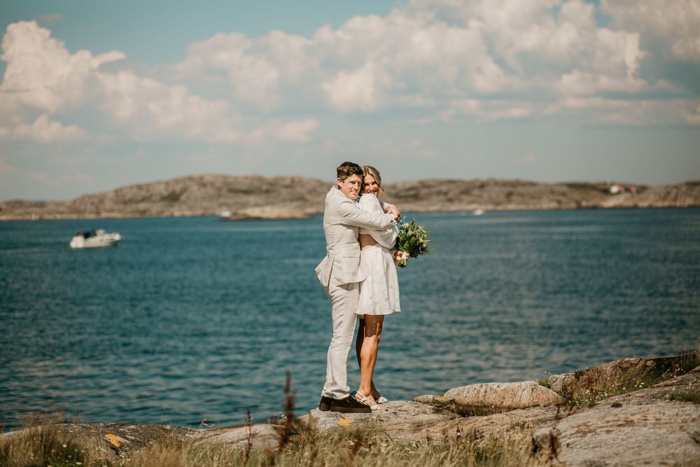 A couple embraces on rocky coastal cliffs overlooking blue waters and distant hills on a sunny summer day.