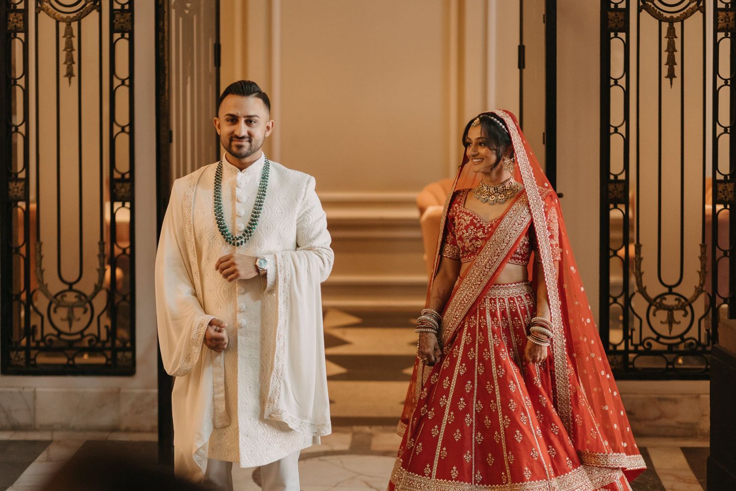 Traditional Indian wedding attire featuring an elegant red lehenga and white sherwani in an ornate venue setting.