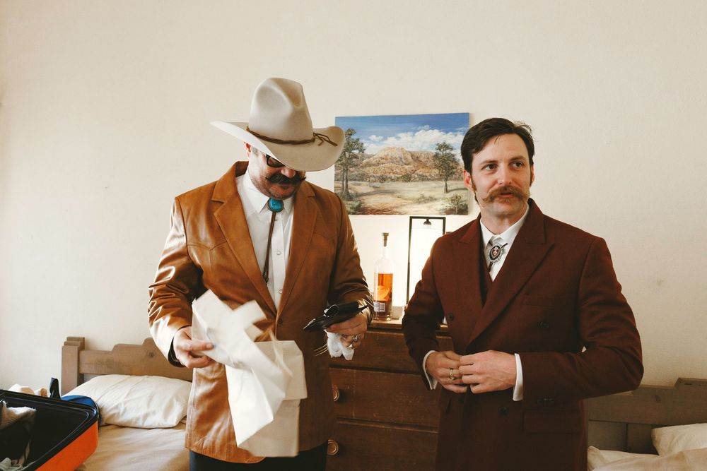 Two groomsmen getting ready indoors, one wearing cowboy hat and brown leather jacket in rustic room.