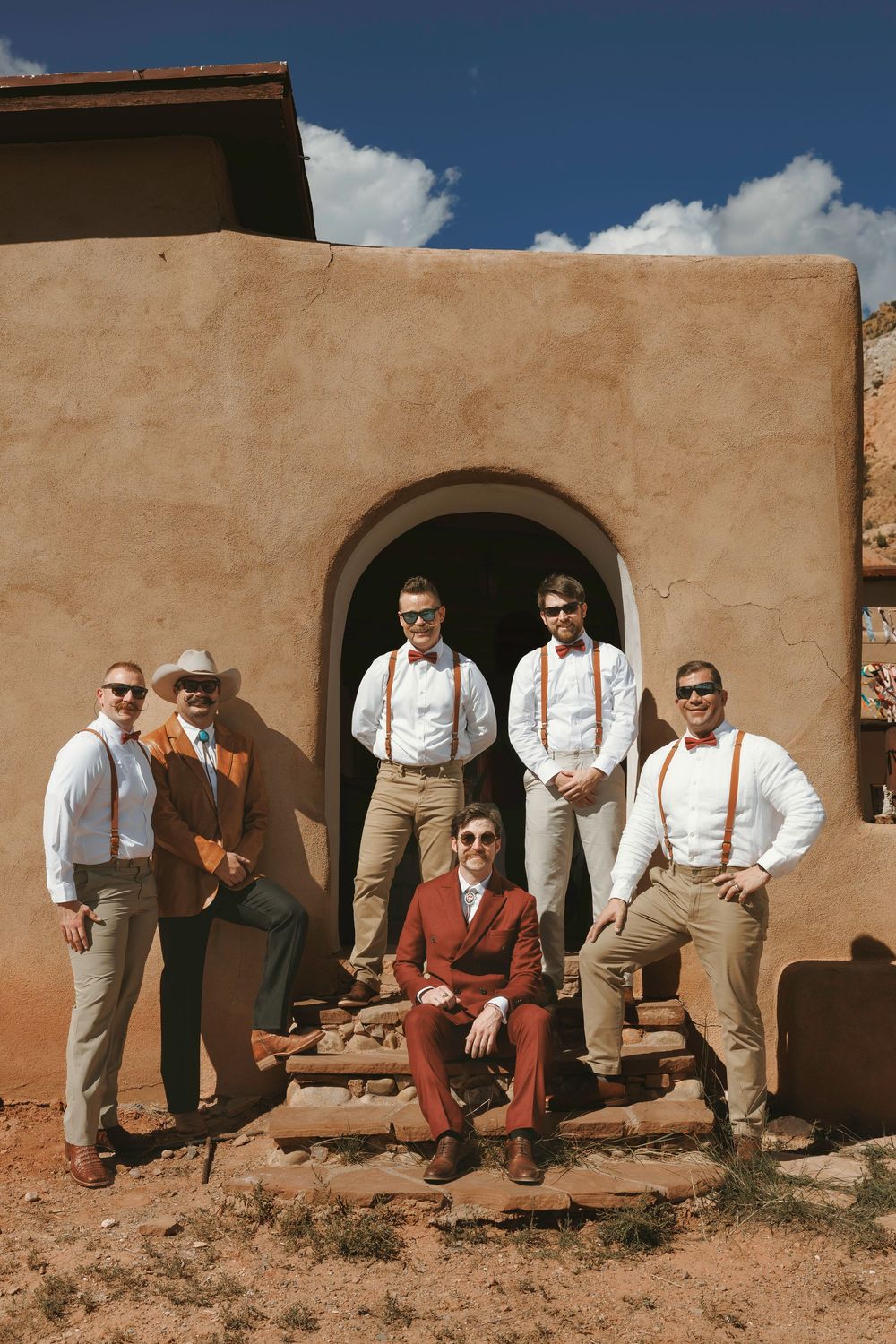 Groom in burgundy suit with groomsmen in white shirts and suspenders posing outside adobe building under blue sky.