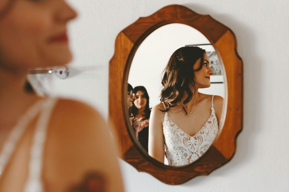 Bride in lace wedding gown smiling in vintage wooden mirror reflection with bridesmaid nearby.