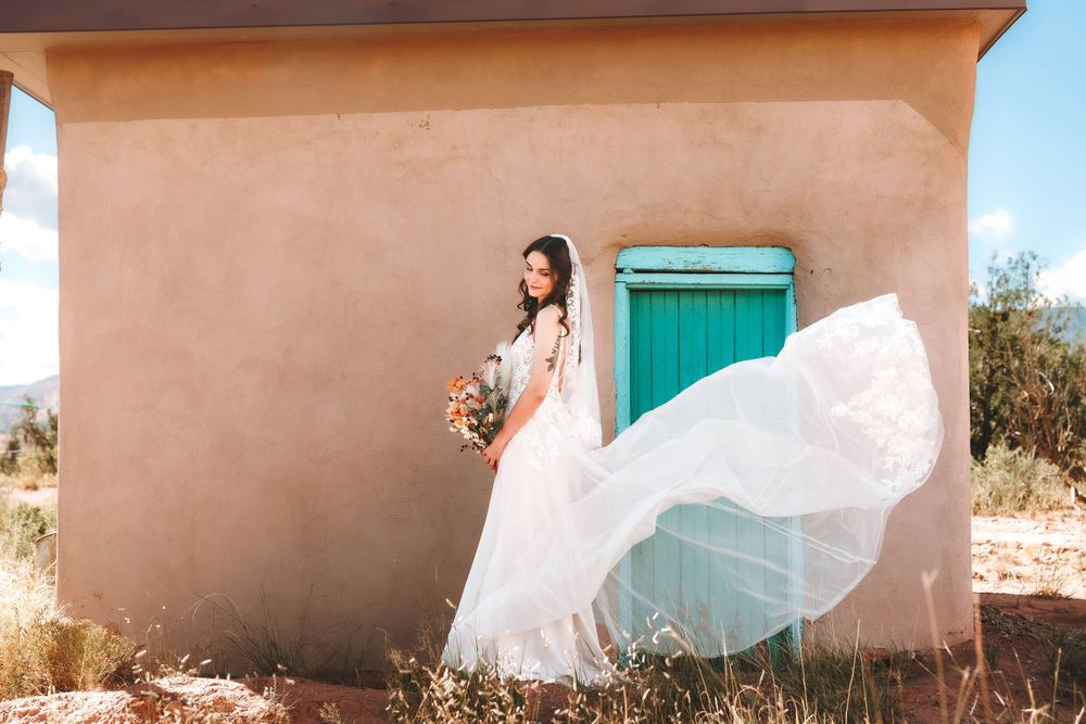 Bride in flowing white gown with veil blowing in wind beside turquoise door on adobe wall.