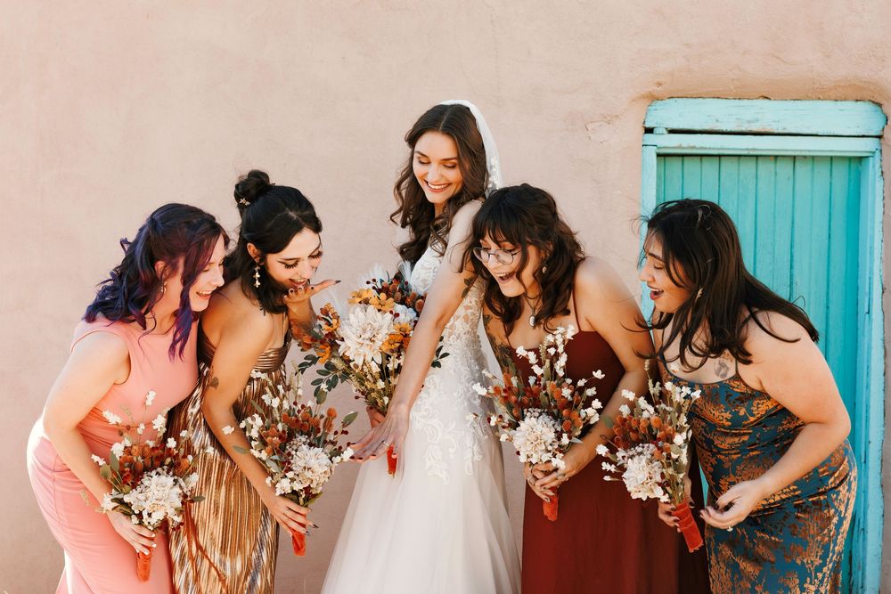 Bride with bridesmaids in mismatched earth-tone dresses holding dried floral bouquets outdoors.