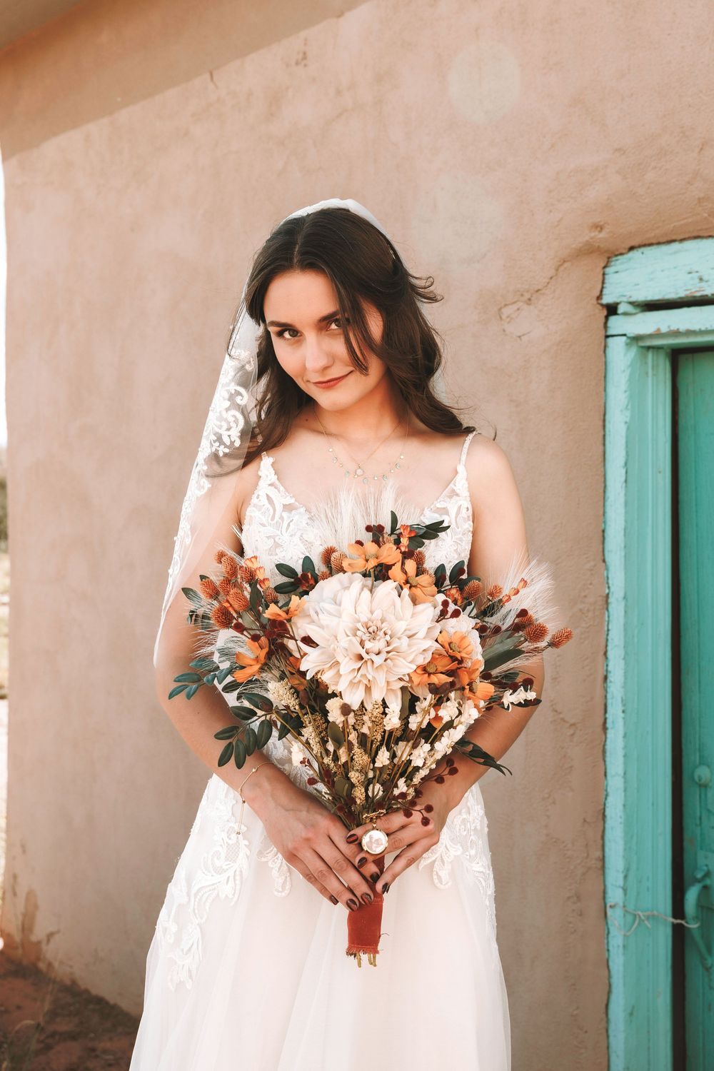 Bride in lace wedding gown holding rustic autumn bouquet with dahlias and dried flowers against adobe wall.