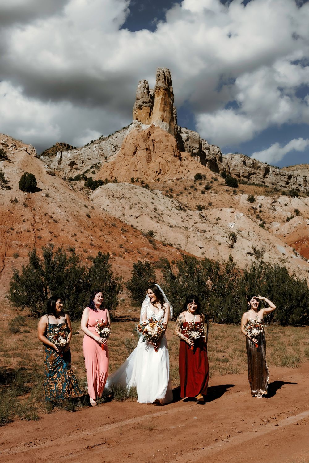 Bride and bridesmaids in colorful mismatched dresses posing on red dirt road with dramatic desert rock formation.