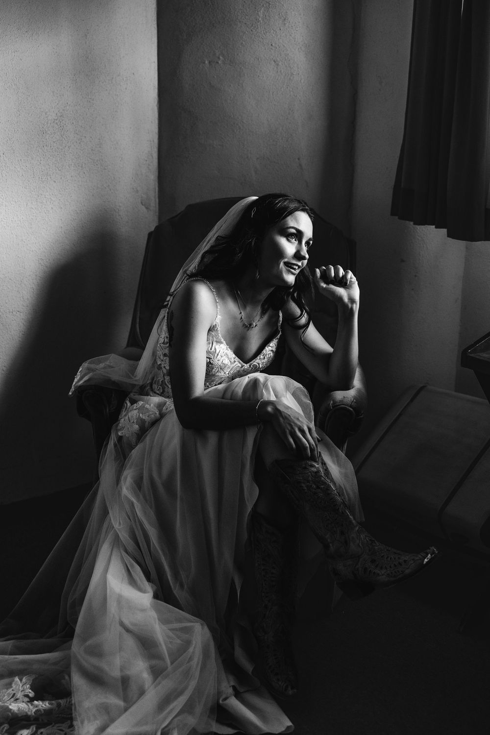 Elegant black and white portrait of smiling bride in lace gown and veil seated in chair by window light.