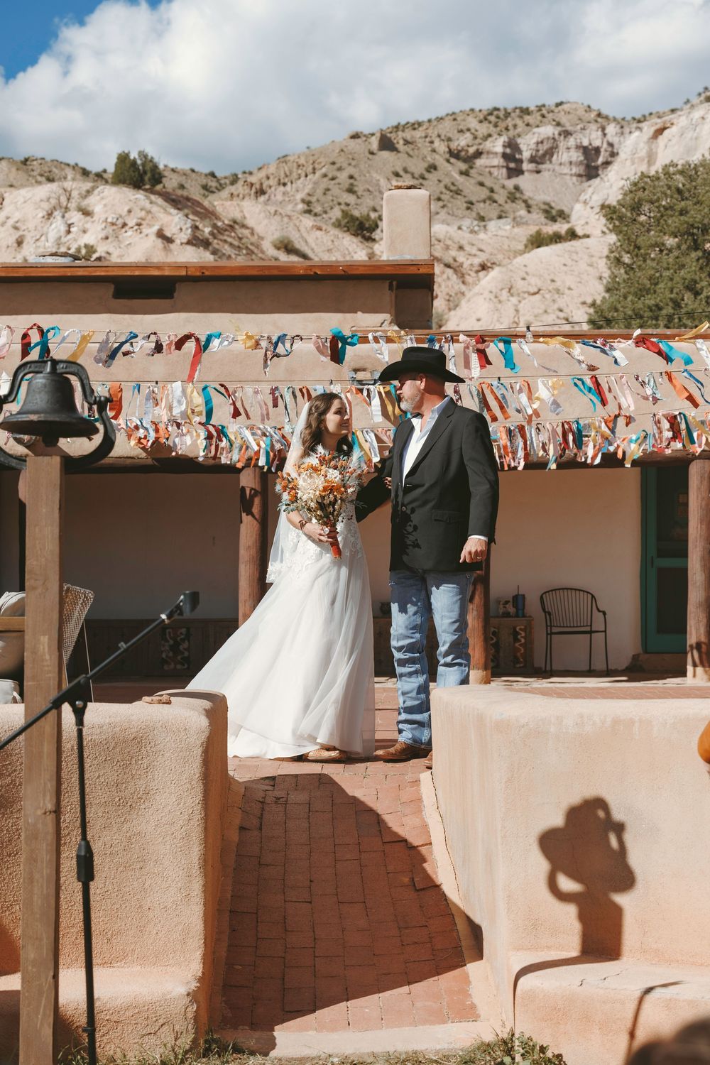 Bride and groom in cowboy hat stand at rustic adobe wedding venue with colorful ribbon decorations.