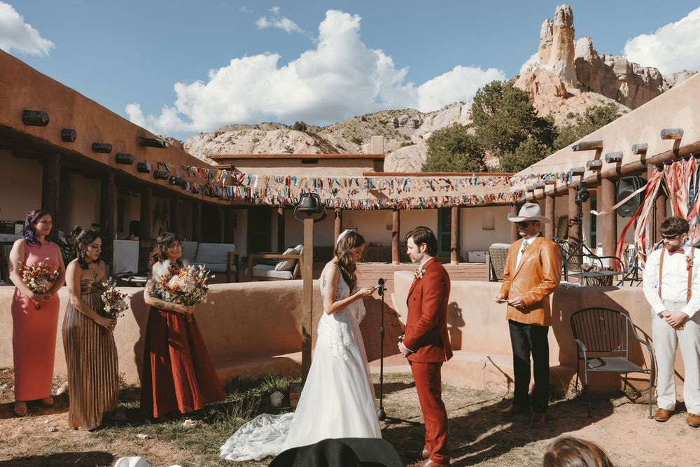 Outdoor wedding ceremony at a southwestern adobe venue with colorful bunting, rocky cliffs, and bridal party in earthy tones.