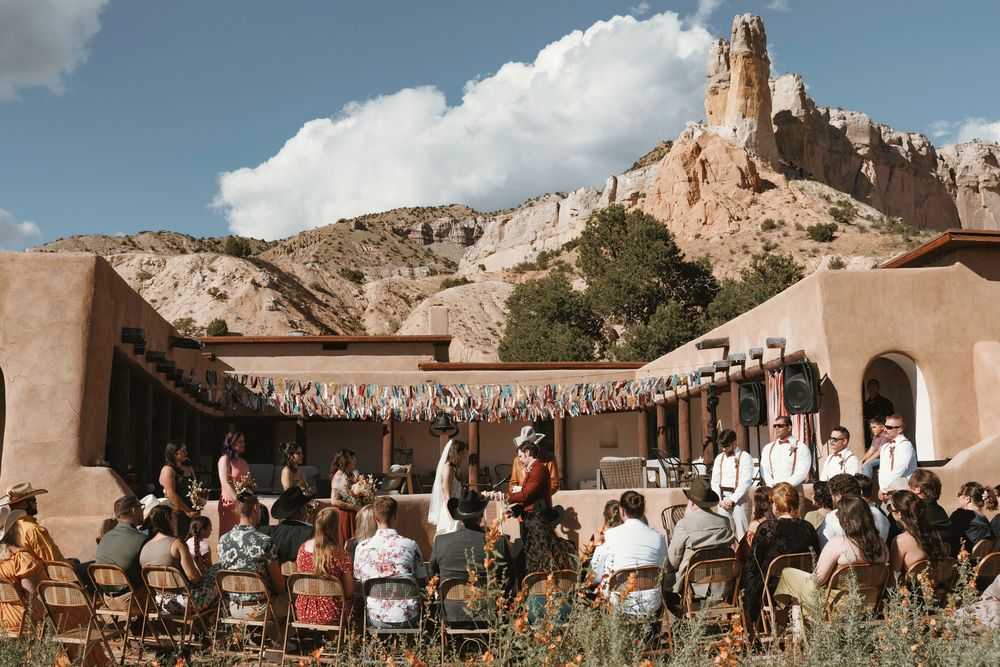 Wedding guests seated outdoors at a desert adobe venue with dramatic rock formations and colorful ribbon garlands overhead.