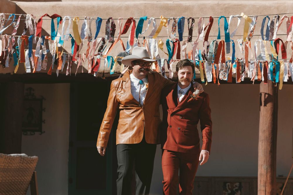 Officiant in tan leather jacket and cowboy hat walks with groom at adobe wedding venue.