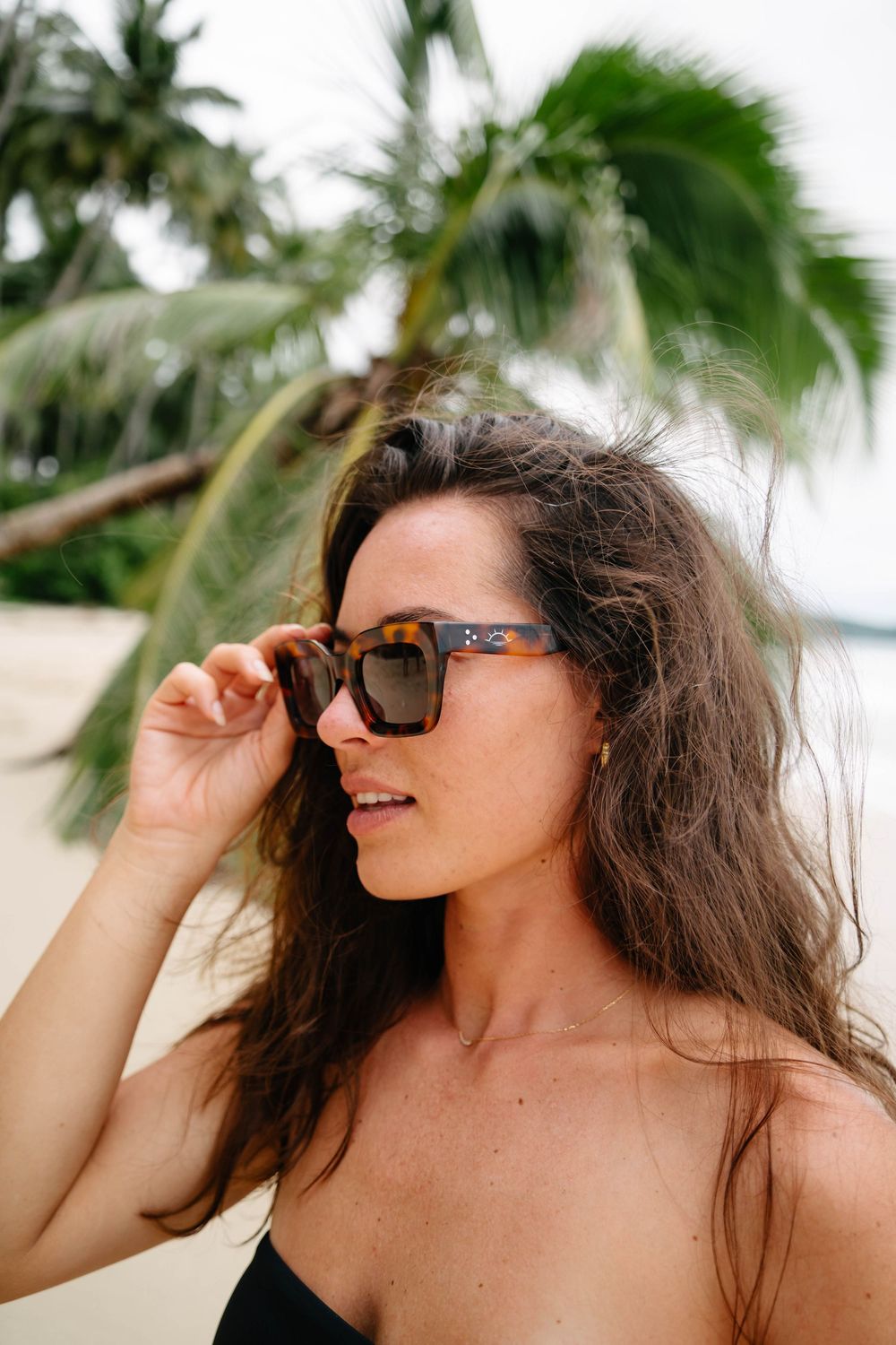 Person wearing sunglasses smiles on tropical beach with palm trees swaying in background.