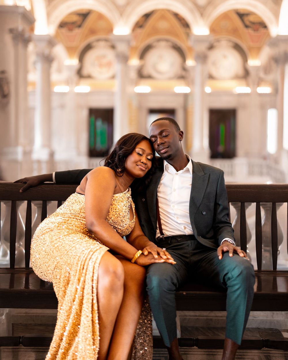 A couple shares intimate moments while sitting on a wooden bench in an ornate indoor setting with arched architecture.