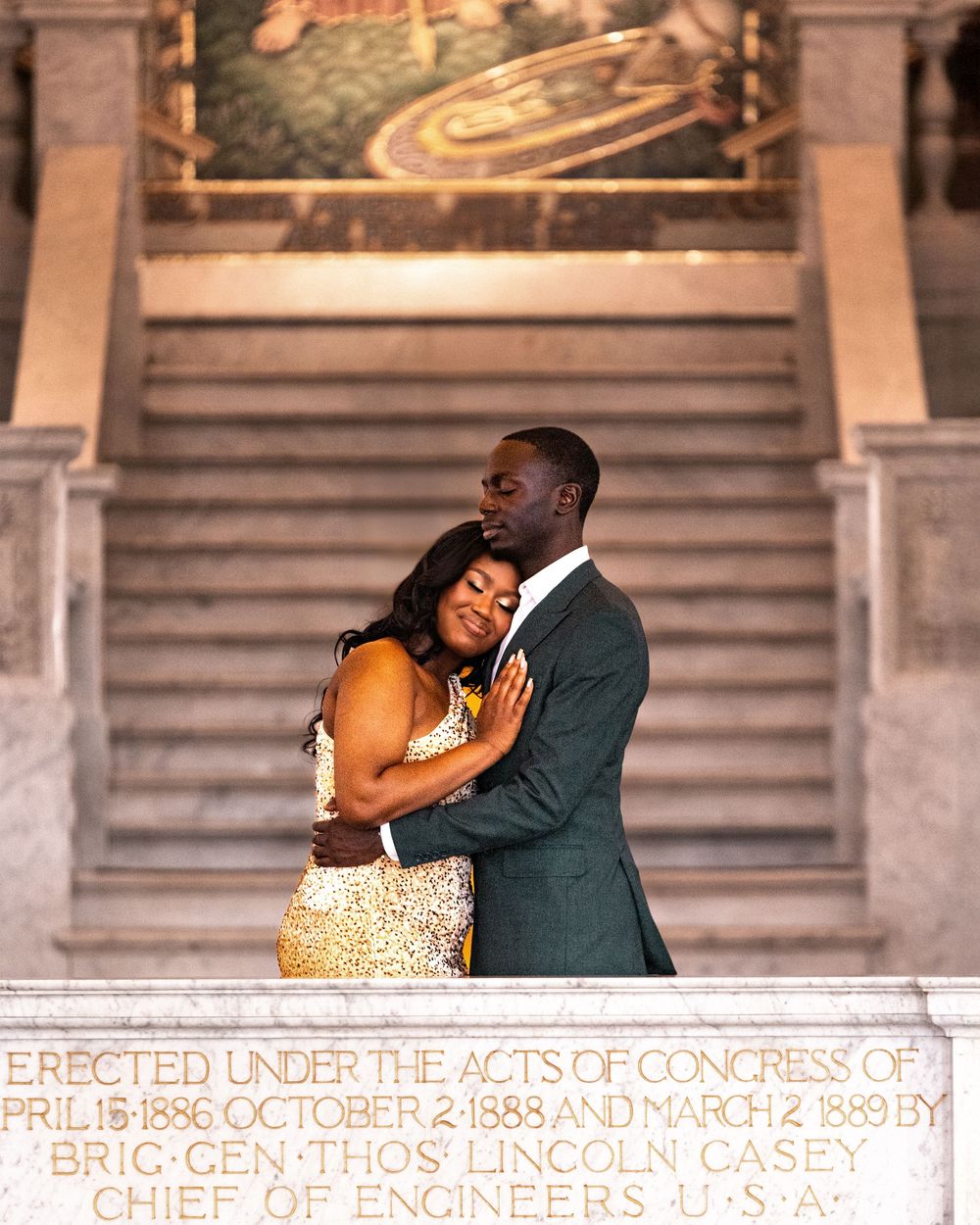 A couple embraces on marble steps in front of historic architectural text engravings.