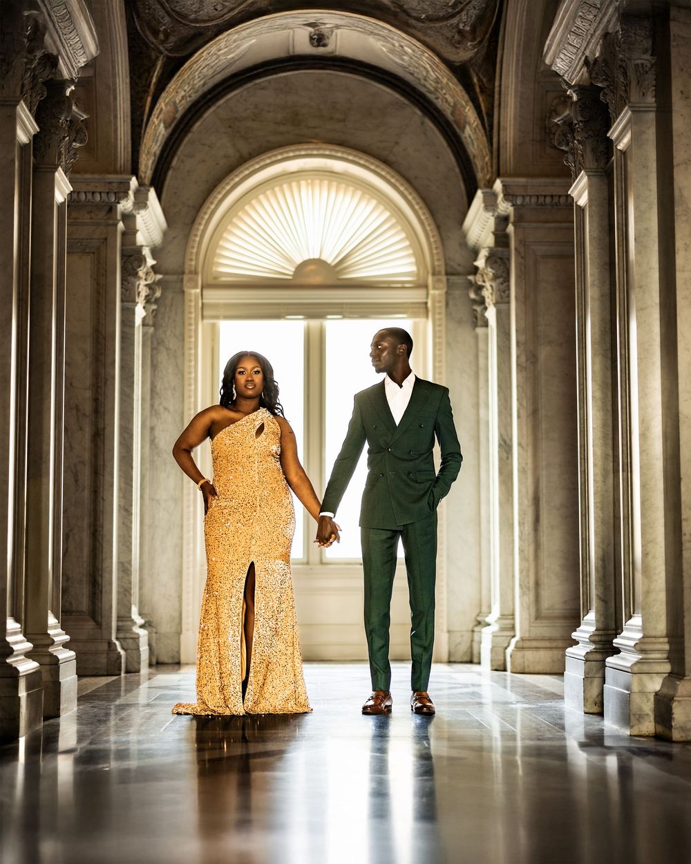 A couple in yellow and green formal attire poses in an elegant marble hallway with arched columns and natural lighting.