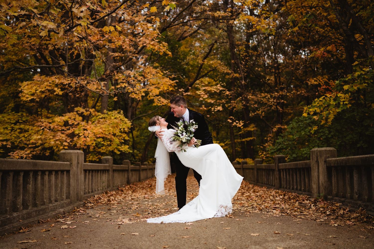 A couple in wedding attire stands on a wooden bridge surrounded by vibrant autumn foliage in a park setting.