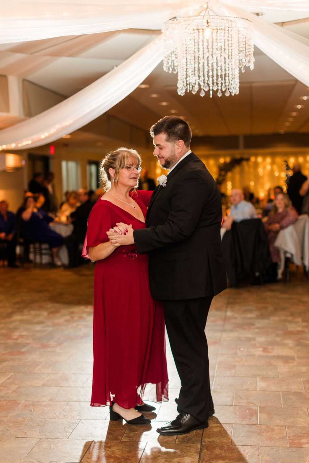A couple in formal attire dancing together at an elegant reception venue with crystal chandeliers overhead.