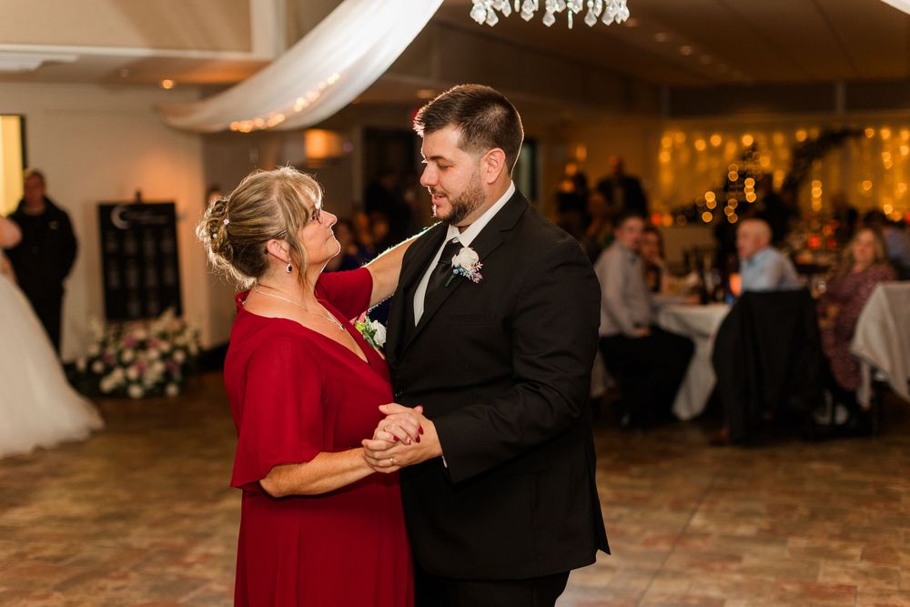 A couple shares an intimate dance moment in formal attire at an indoor venue with warm lighting and holiday decor.