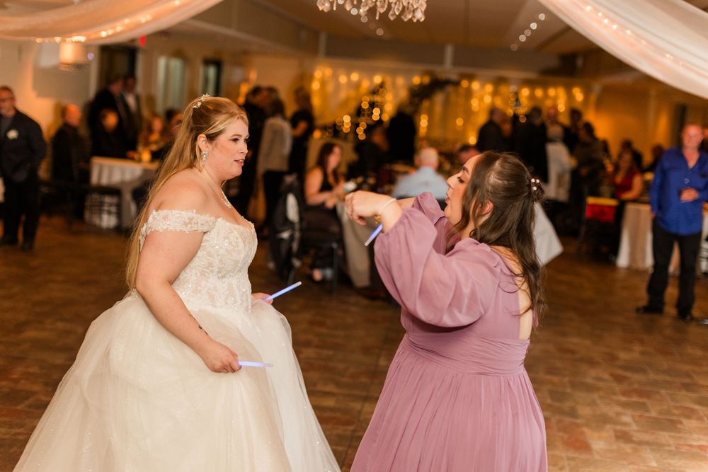 Wedding party dances together on a wooden dance floor in a reception hall with twinkling lights overhead.