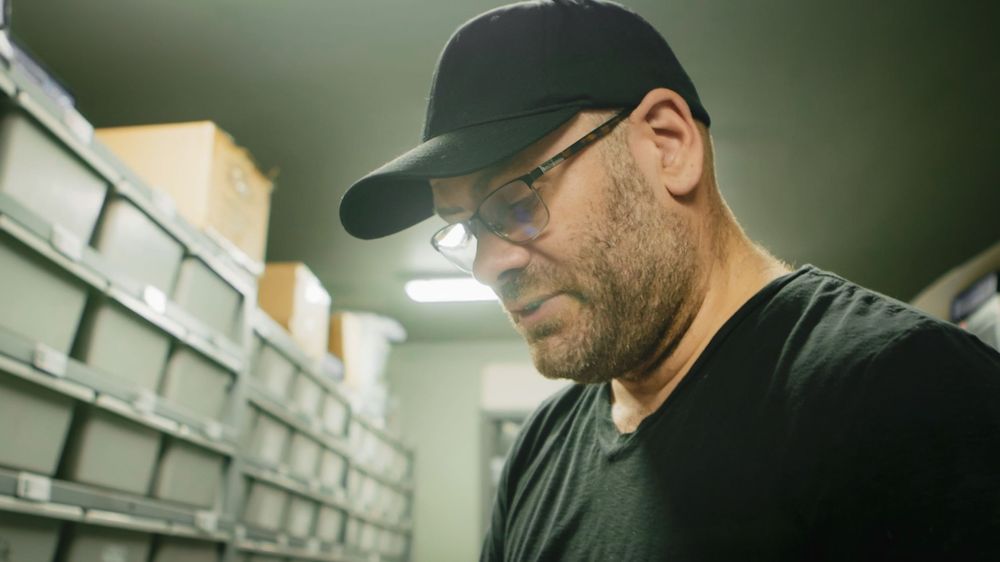 Person wearing a black baseball cap and dark shirt examining items on shelves in a dimly lit storage area.