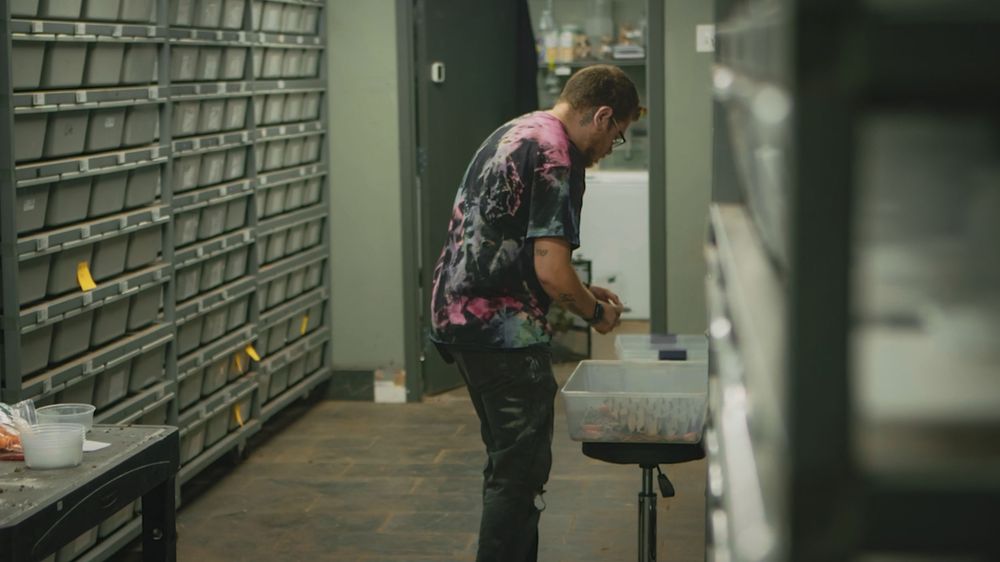 Warehouse worker in dark uniform moves between tall storage shelves filled with packages and boxes.
