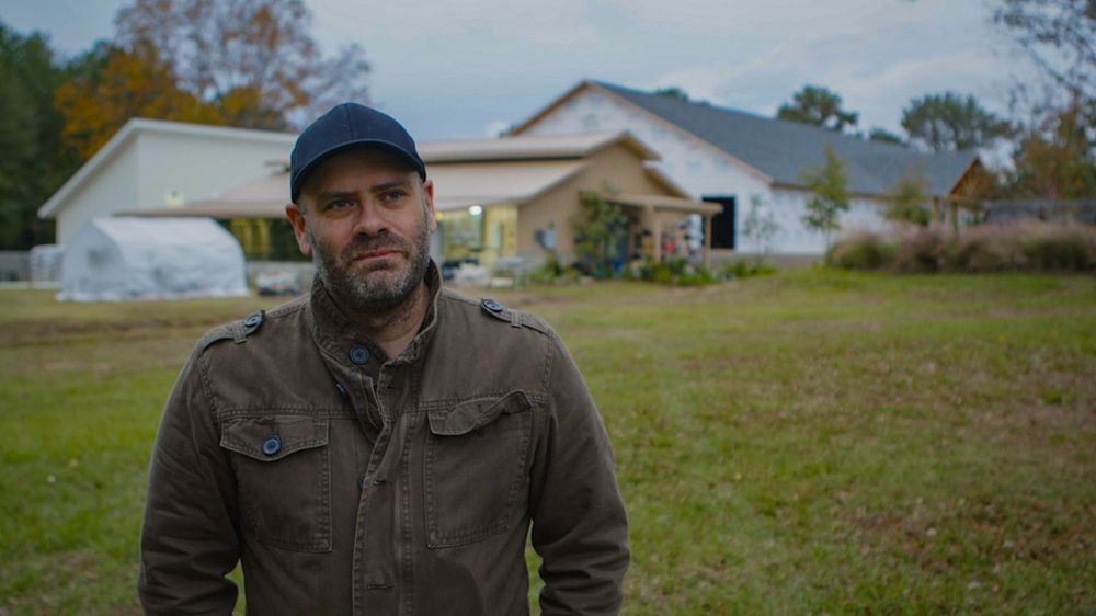 A person wearing a baseball cap and brown jacket stands in front of a rural farmhouse on a grassy field.