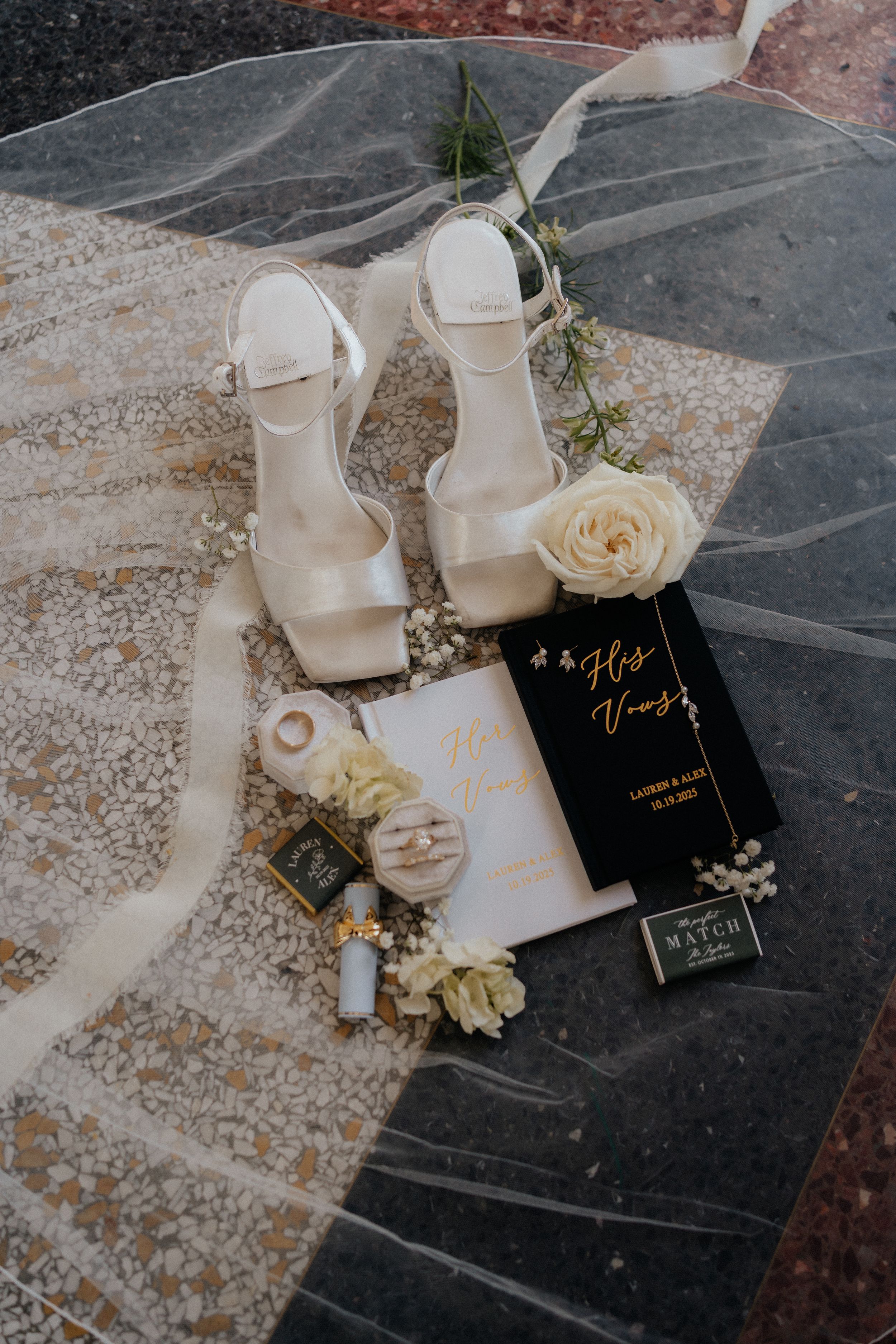Elegant white wedding shoes with roses and vow books on a marble surface create a romantic bridal flatlay.