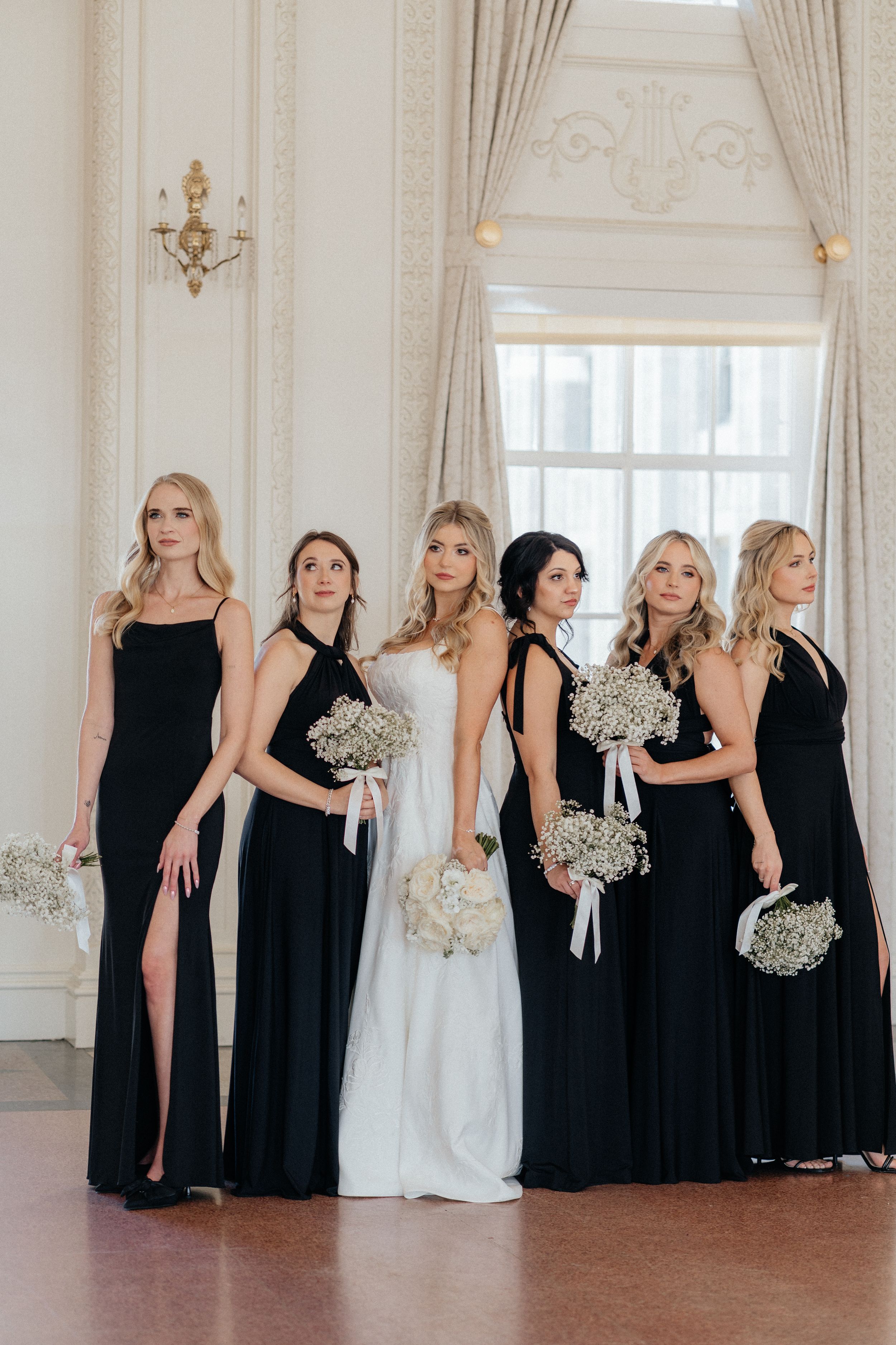 Bridesmaids in elegant black dresses pose with white bouquets in an ornate white ballroom.