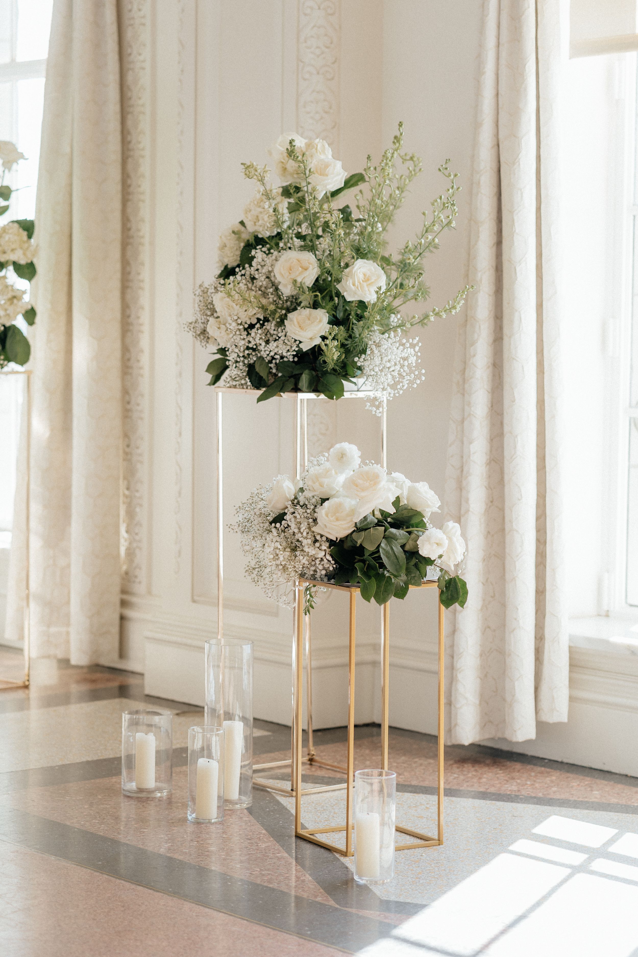 Modern white wedding ceremony space with tall floral arrangements on gold stands near sunlit windows.