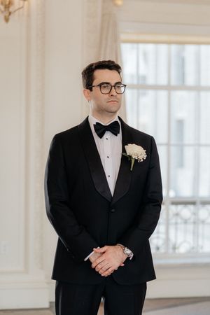 A groom in a classic black tuxedo and bow tie poses in an elegant white room.