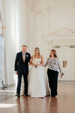 A wedding party walks through an elegant white ballroom with ornate moldings and architectural details.