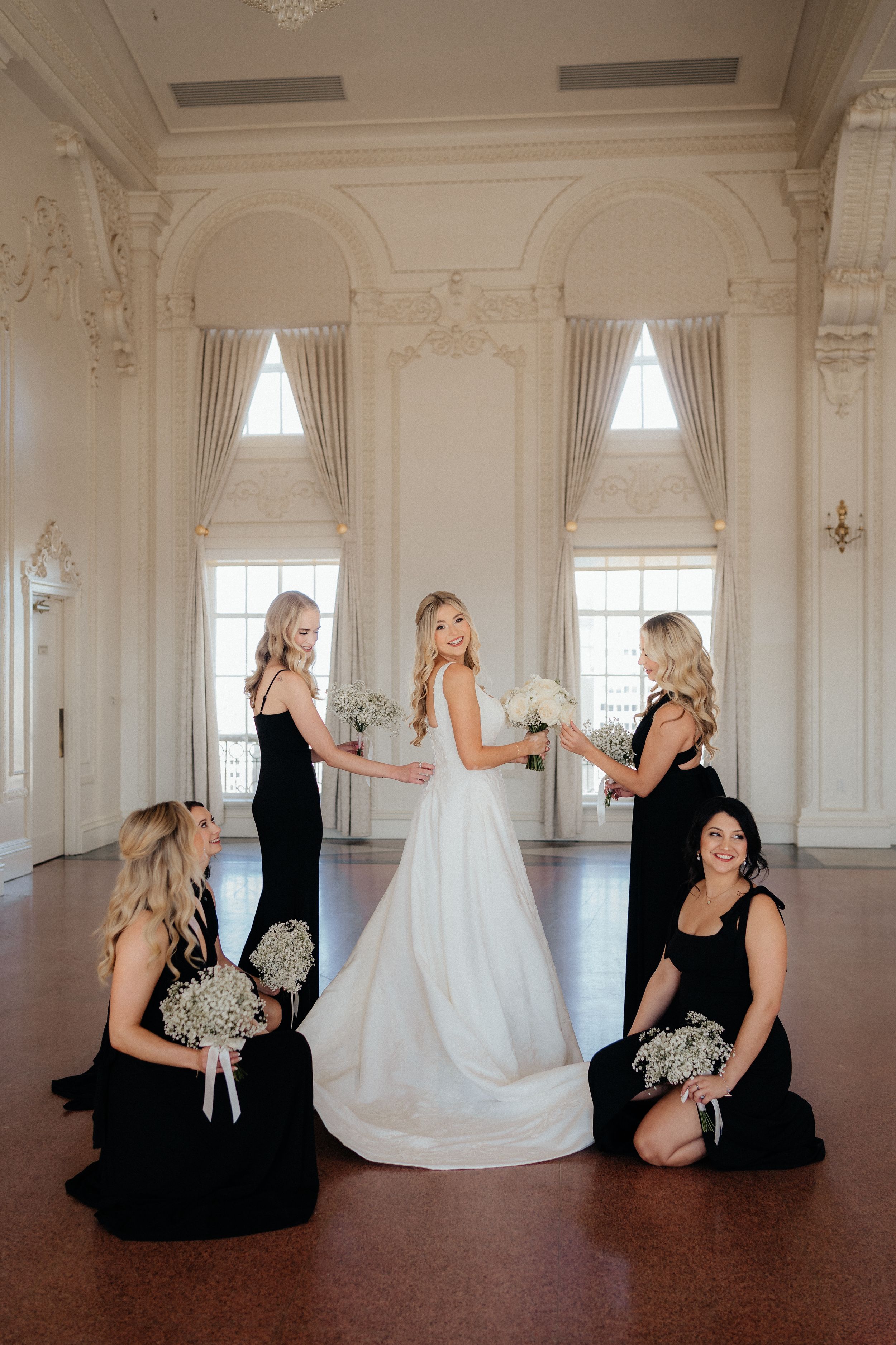 Wedding party in black dresses and white gown posing playfully in an ornate white ballroom.
