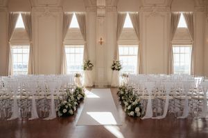 Elegant white wedding venue with high ceilings, ornate moldings, and rows of white chairs arranged for a ceremony.