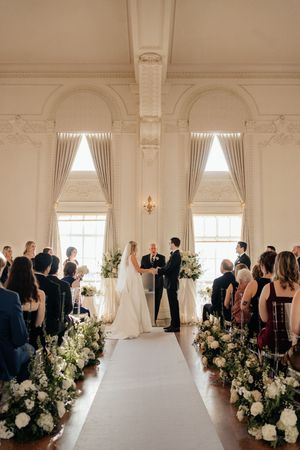 Wedding ceremony in a bright white room with floral arrangements lining the aisle and guests seated.